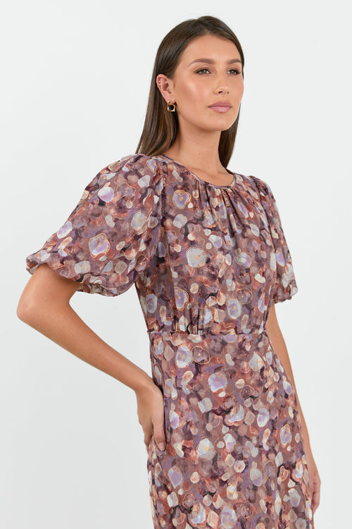 A woman wearing a floral midi dress in shades of lilac, blush, and soft pink, with puffed short sleeves and a relaxed fit.