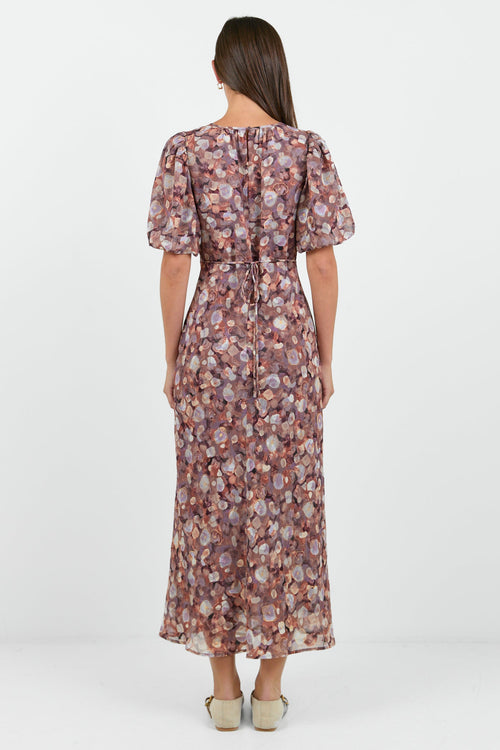 A woman wearing a floral midi dress in shades of lilac, blush, and soft pink, with puffed short sleeves and a relaxed fit.