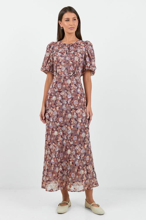 A woman wearing a floral midi dress in shades of lilac, blush, and soft pink, with puffed short sleeves and a relaxed fit.