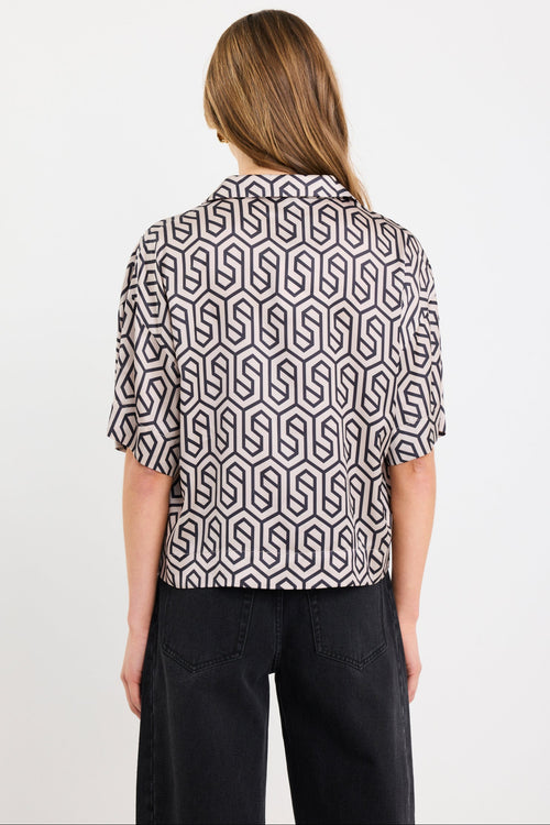 Woman wearing a short sleeve satin shirt with bold black geometric print, button front, and tie waist detail, styled with black wide-leg jeans and white sandals.