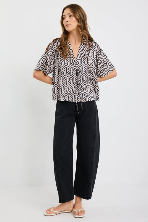 Woman wearing a short sleeve satin shirt with bold black geometric print, button front, and tie waist detail, styled with black wide-leg jeans and white sandals.