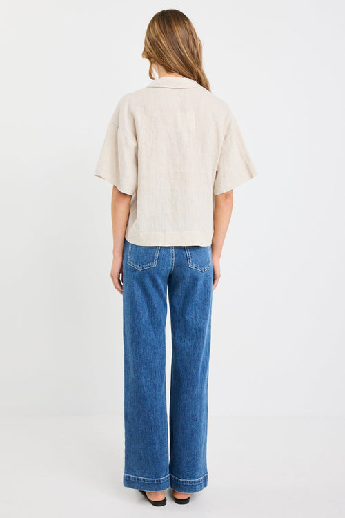 Woman wearing a natural beige short sleeve linen shirt with tie-front fastening, styled with mid-blue jeans.