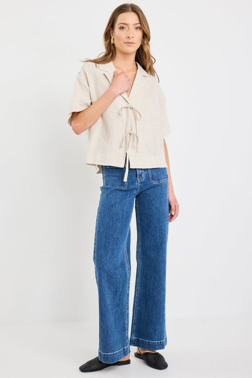 Woman wearing a natural beige short sleeve linen shirt with tie-front fastening, styled with mid-blue jeans.