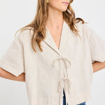 Woman wearing a natural beige short sleeve linen shirt with tie-front fastening, styled with mid-blue jeans.