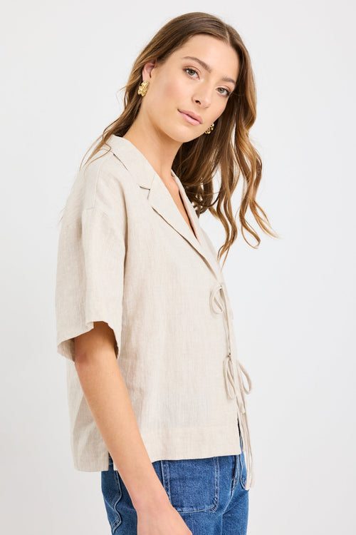 Woman wearing a natural beige short sleeve linen shirt with tie-front fastening, styled with mid-blue jeans.