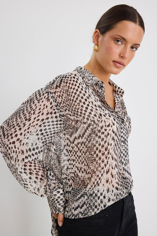 Woman wearing a loose-fitting, long-sleeve button-up shirt in a charcoal snake print, styled half-tucked into black jeans with gold hoop earrings.