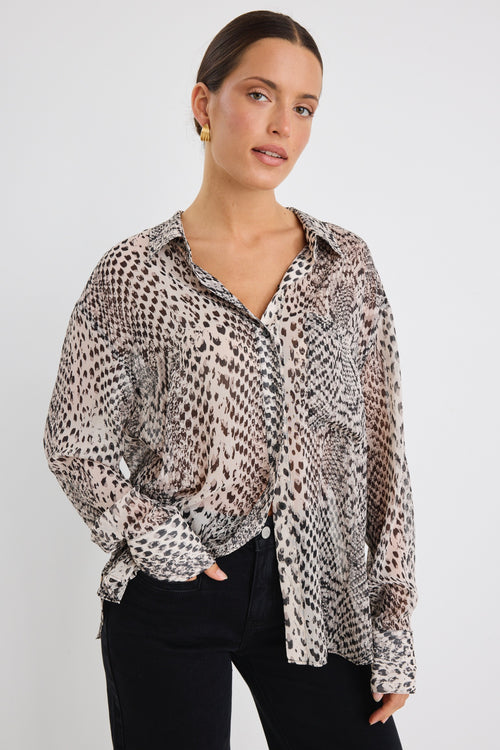 Woman wearing a loose-fitting, long-sleeve button-up shirt in a charcoal snake print, styled half-tucked into black jeans with gold hoop earrings.