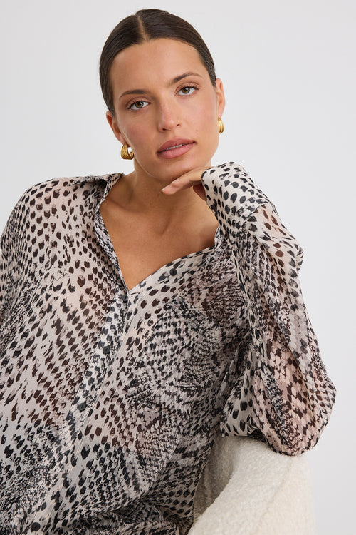 Woman wearing a loose-fitting, long-sleeve button-up shirt in a charcoal snake print, styled half-tucked into black jeans with gold hoop earrings.