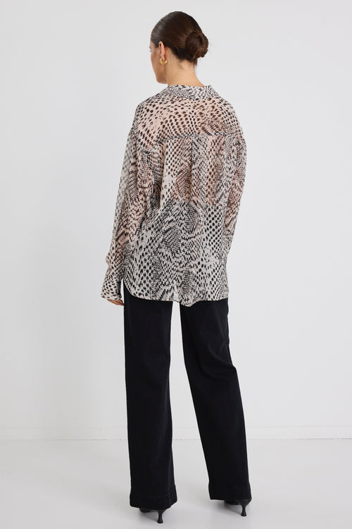 Woman wearing a loose-fitting, long-sleeve button-up shirt in a charcoal snake print, styled half-tucked into black jeans with gold hoop earrings.