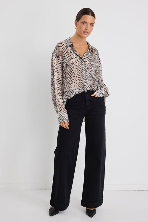 Woman wearing a loose-fitting, long-sleeve button-up shirt in a charcoal snake print, styled half-tucked into black jeans with gold hoop earrings.