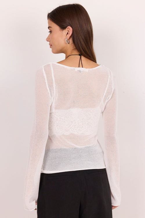 A woman shown from the back wearing a sheer white long-sleeve knit top with a lace panel and black pants, against a plain off-white studio background.