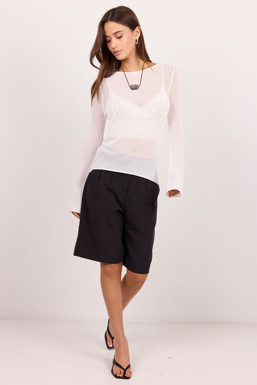 A woman modeling a sheer white long-sleeve top over a white lace camisole, paired with black shorts and black strappy heels, against a plain white studio background.
