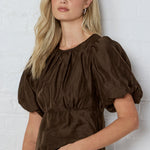 Woman wearing a brown blouse with puffed sleeves against a white brick wall.
