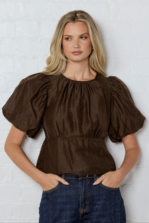 Woman wearing a brown blouse with puffed sleeves against a white wall