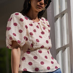 A woman wearing a cream puff-sleeve top with red polka dots and blue jeans, standing on a balcony with white-framed windows.
