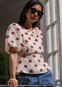 A woman wearing a cream puff-sleeve top with red polka dots and blue jeans, standing on a balcony with white-framed windows.