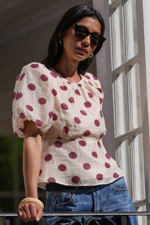 A woman wearing a cream puff-sleeve top with red polka dots and blue jeans, standing on a balcony with white-framed windows.