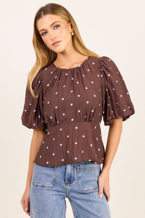 A woman modeling a brown short-sleeve top with white polka dots and puffed sleeves, paired with light blue jeans, against a plain cream background.