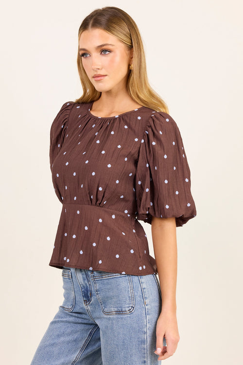 A woman modeling a brown short-sleeve blouse with white polka dots and puffed sleeves, paired with light blue jeans, against a plain cream background.