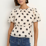 Woman wearing a cream short sleeve top with black polka dots, puff sleeves, and blue jeans.