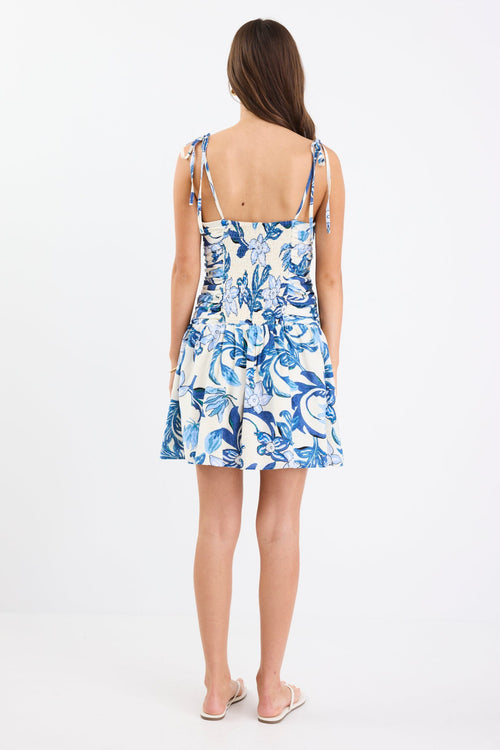 Woman wearing a blue floral mini dress with a pleated bodice, strappy sleeves, and a flattering fit, paired with white sandals.