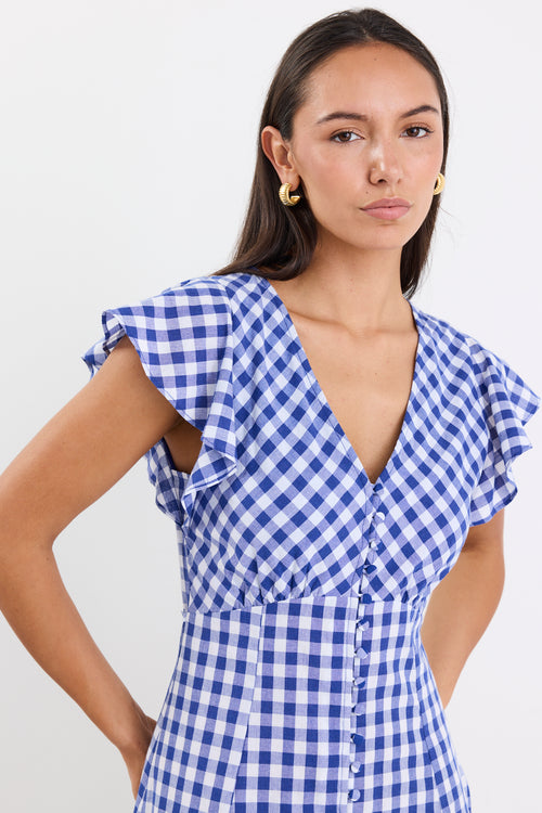Woman wearing a blue gingham midi dress with a V-neck, button-down front, and fluted sleeves, paired with nude sandals.