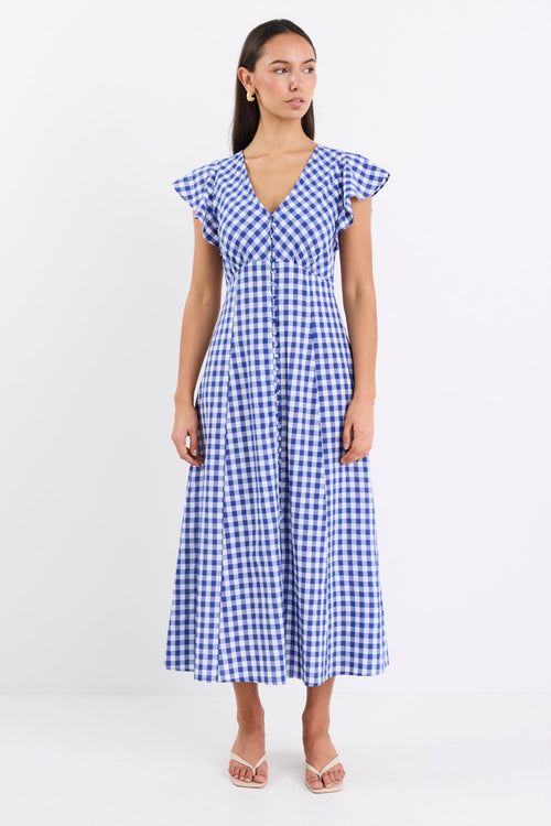 Woman wearing a blue gingham midi dress with a V-neck, button-down front, and fluted sleeves, paired with nude sandals.