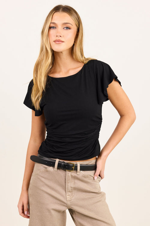 A blonde woman modeling a black short-sleeve top with ruching, paired with beige pants and a black belt, against a plain off-white studio background.