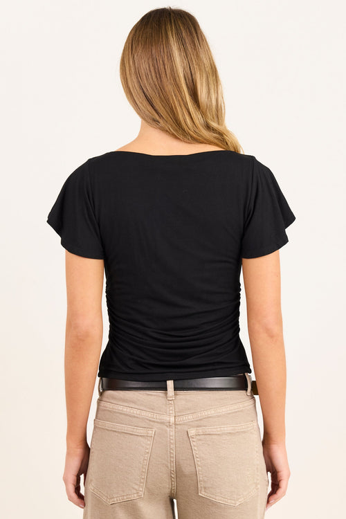 A woman viewed from behind wearing a black short-sleeve top with a wide boat neckline and beige pants, against a plain off-white background.
