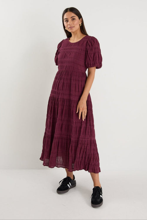 Woman wearing a plum puff-sleeve maxi dress with a textured finish and tiered skirt, paired with black sneakers.