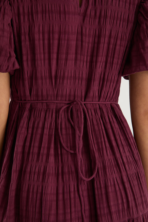 Woman wearing a plum puff-sleeve maxi dress with a textured finish and tiered skirt, paired with black sneakers.