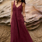 A woman wearing a sleeveless, V-neck maxi dress in deep burgundy stands on a rock in front of a large layered sandstone cliff.