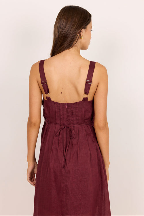 A woman shown from the back wearing a sleeveless burgundy midi dress with adjustable shoulder straps and a tie at the waist, photographed against a plain light background.