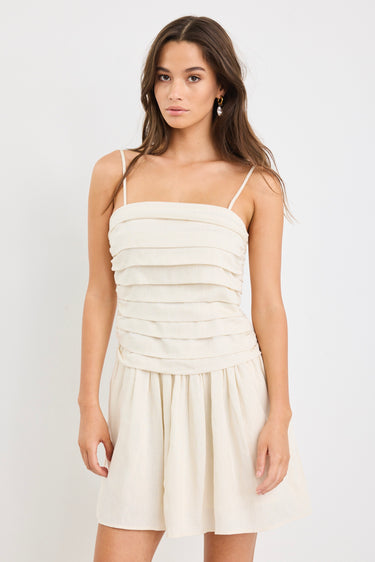 Woman wearing a plain ecru mini dress with thin spaghetti straps, a pleated bodice, and softly flared skirt.