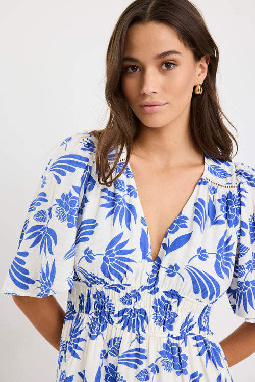 Woman wearing a white maxi dress with bold blue floral print, flutter sleeves, v-neckline, and tiered skirt, styled with neutral sandals.