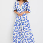 Woman wearing a white maxi dress with bold blue floral print, flutter sleeves, v-neckline, and tiered skirt, styled with neutral sandals.