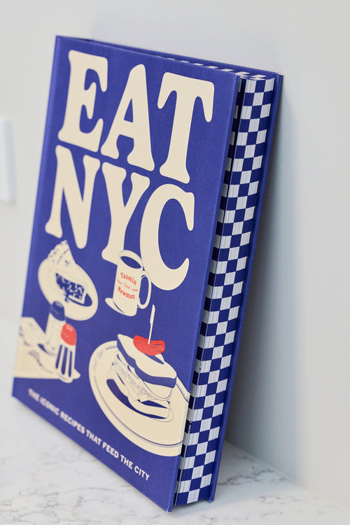 A hardcover book titled 'EAT NYC' leaning against a white wall on a light marble surface. The cover is predominantly blue with large cream lettering and illustrated food and drink graphics, and the spine shows a blue-and-white checkered pattern.