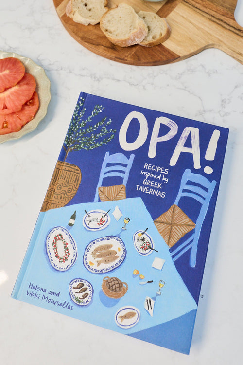 A cookbook titled 'OPA! Recipes for a Better Gathering' placed on a white marble countertop next to a charcuterie board with tomatoes, bread, olives, and cheese, with a plate of sliced tomato and cheese nearby.
