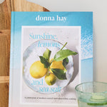 A book titled "Sunshine Lemon and Lime" by donna hay, standing upright on a white marble countertop next to a wooden cutting board, a small green-tinted glass, and a plate of lemons against a white wall.