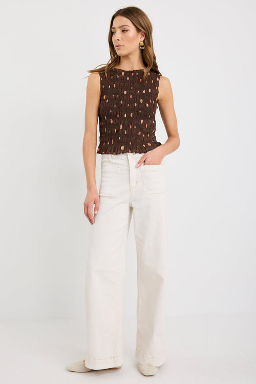 Woman wearing a sleeveless chocolate brown shirred top with scattered polka dot print and frilled hem, styled with ecru jeans.