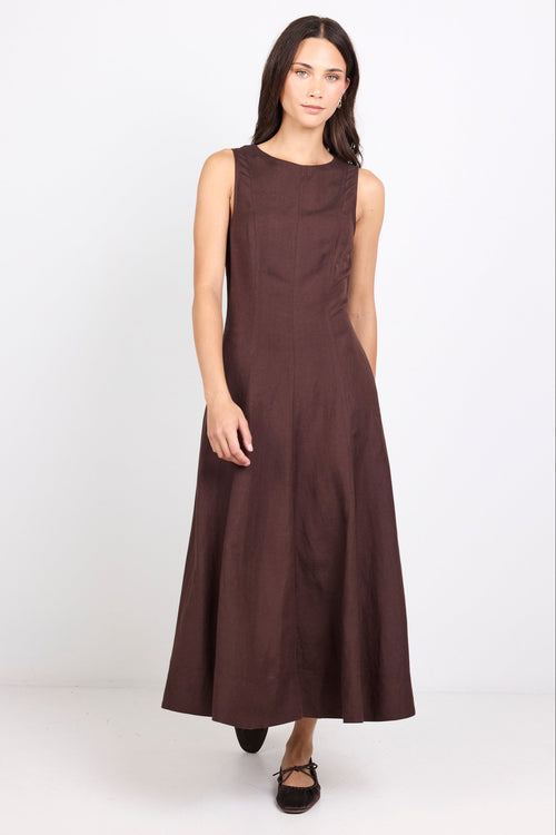 A woman modeling a sleeveless, dark brown midi dress with a high round neckline and a flowing skirt, standing against a plain white studio background.