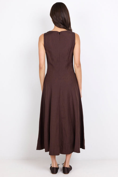 A woman viewed from the back wearing a sleeveless dark brown midi dress and black shoes, standing against a plain white studio background.