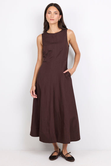 A woman modeling a sleeveless, dark brown midi dress with a high neckline and side pockets, paired with dark flat shoes, against a plain white studio background.