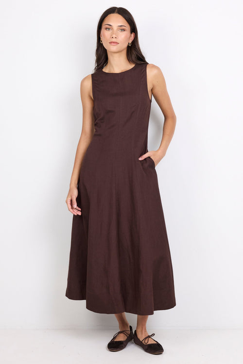 A woman modeling a sleeveless, dark brown midi dress with a high neckline and side pockets, paired with dark flat shoes, against a plain white studio background.