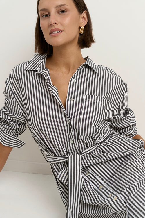 Woman wearing a black and white striped button-down midi shirt dress with a waist tie, rolled sleeves, and white sneakers.