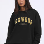 Woman wearing a black crewneck jumper with “FOXWOOD” in metallic gold across the chest, styled casually with relaxed black pants.