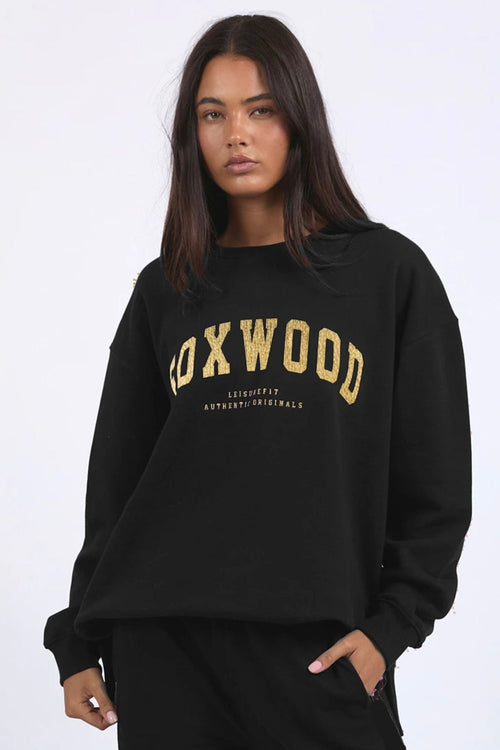 Woman wearing a black crewneck jumper with “FOXWOOD” in metallic gold across the chest, styled casually with relaxed black pants.