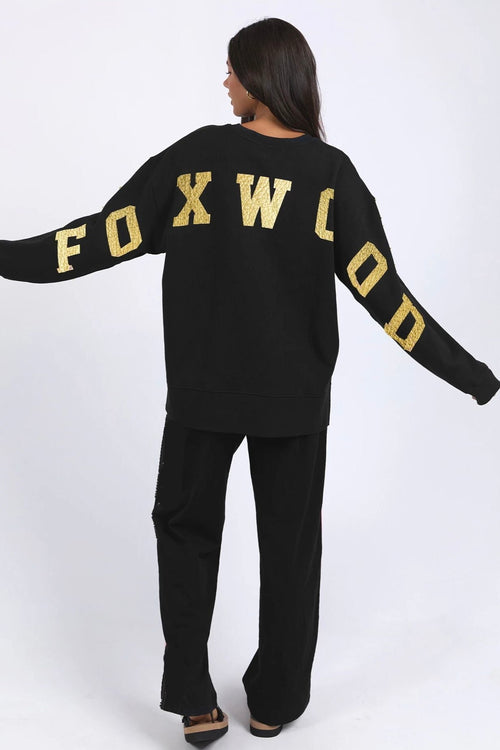 Woman wearing a black crewneck jumper with “FOXWOOD” in metallic gold across the chest, styled casually with relaxed black pants.