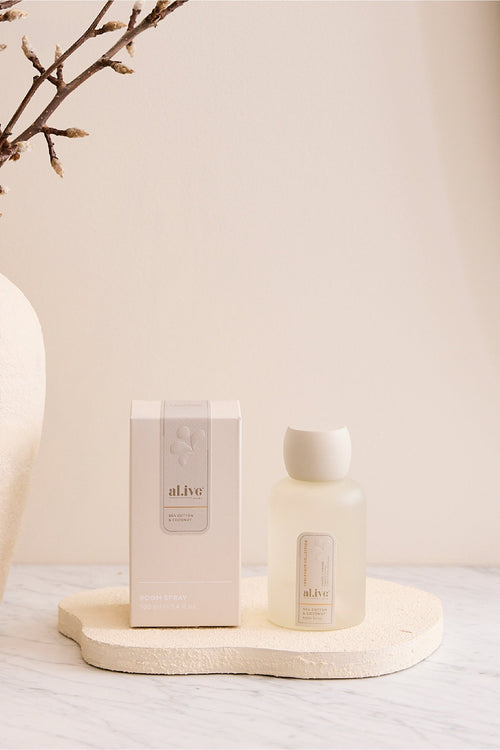 White candle and packaging on a wooden tray with a vase and branches in the background.