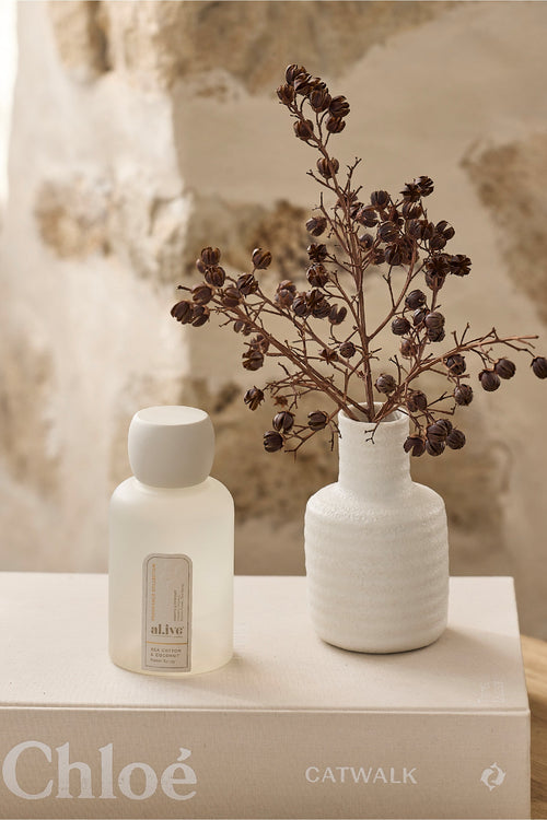 White bottle and vase with dried flowers on a book titled 'Chloe Catwalk' with a textured beige background.